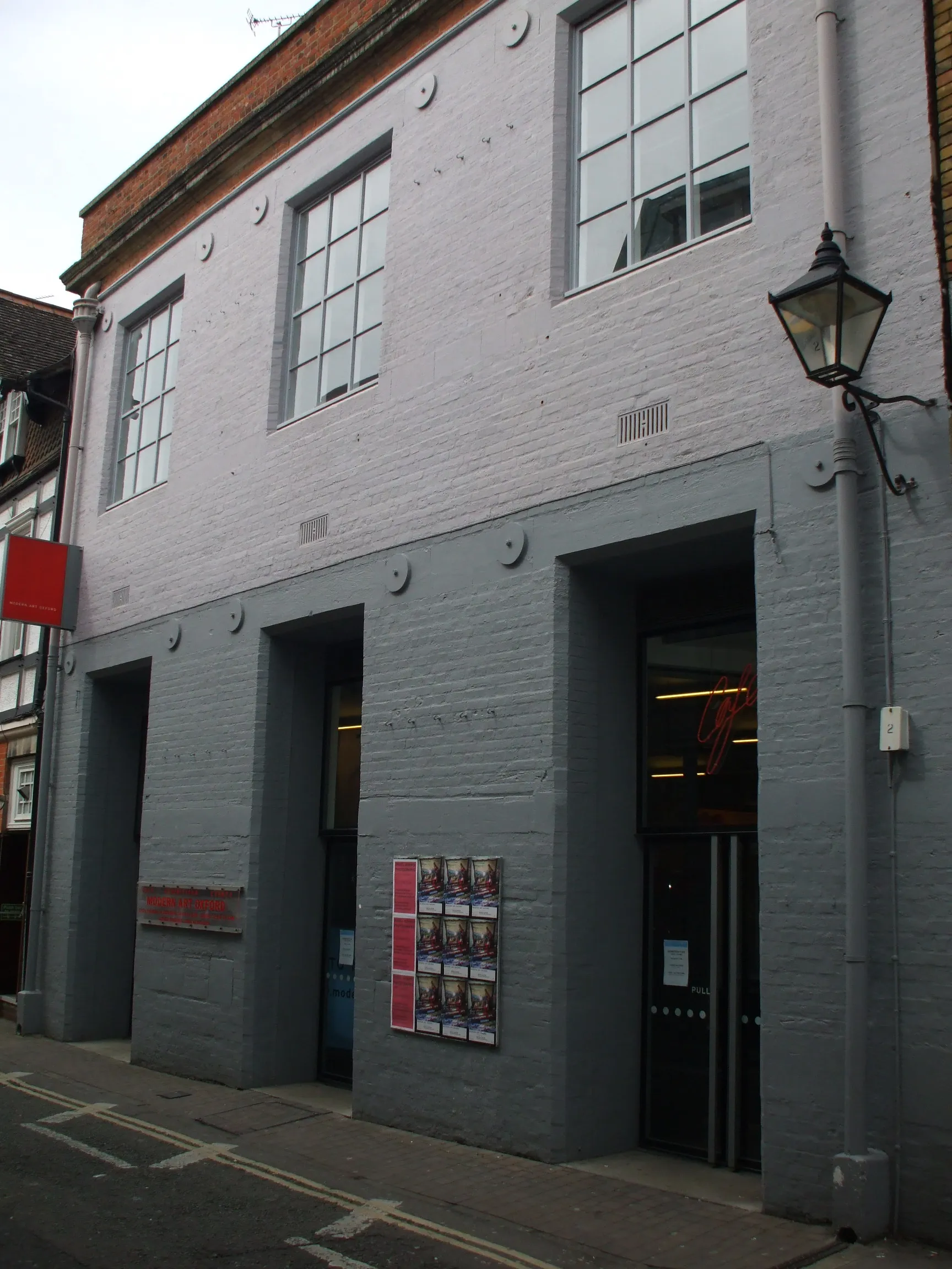 Exterior view of Modern Art Oxford showing a gray painted brick building with large multi-pane windows on the upper floor, three recessed entrance doorways, a vintage street lamp, and exhibition posters on the wall.