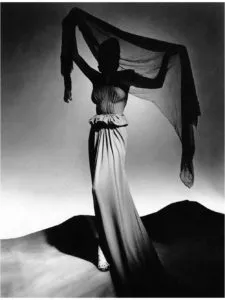 Fotografía de moda en blanco y negro de una figura vista desde atrás sosteniendo una tela oscura y transparente sobre dunas de arena.