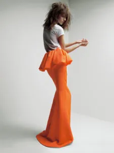 Retrato de cuerpo completo de una modelo que viste una camiseta gris y una falda larga de color naranja brillante con una cintura peplum voluminosa sobre un fondo blanco.