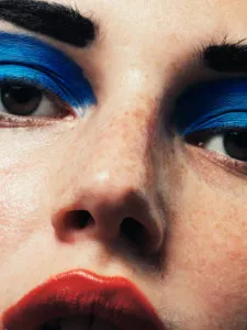 Primer plano extremo del rostro de una persona que luce una atrevida sombra de ojos azul eléctrico, cejas negras gruesas, piel con pecas y labios de un rojo intenso.