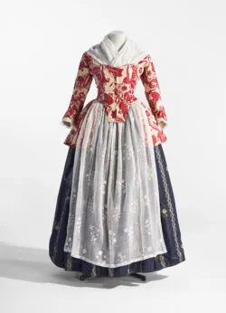 Vestido histórico con cuerpo bordado en rojo y falda plisada de color gris plateado con enagua azul oscuro, exhibido en un maniquí sobre fondo blanco.