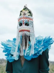 Persoon die een uitbundig artistiek masker draagt met witte franjes, decoratief kralenwerk en een dramatische blauwe veren kraag tegen een bewolkte lucht