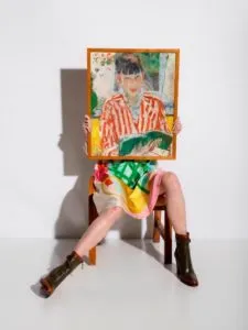 Persona sentada en una silla de madera sosteniendo un cuadro enmarcado frente a la cara, vistiendo una camiseta de rayas rojas, falda colorida y botines marrones sobre un fondo blanco