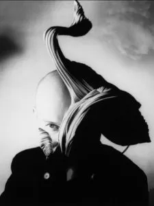 Fotografía en blanco y negro de una figura calva con un elemento de tela escultórico y dramático que se eleva desde los hombros, con la mano apoyada cerca del rostro en una pose contemplativa.