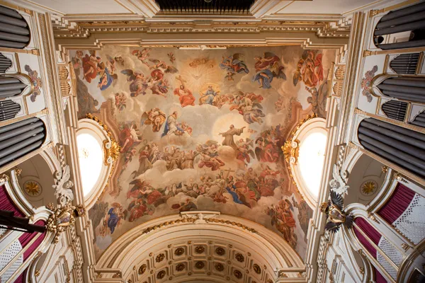 Ornate ceiling fresco with religious figures in a grand interior space, flanked by large organs and arched windows