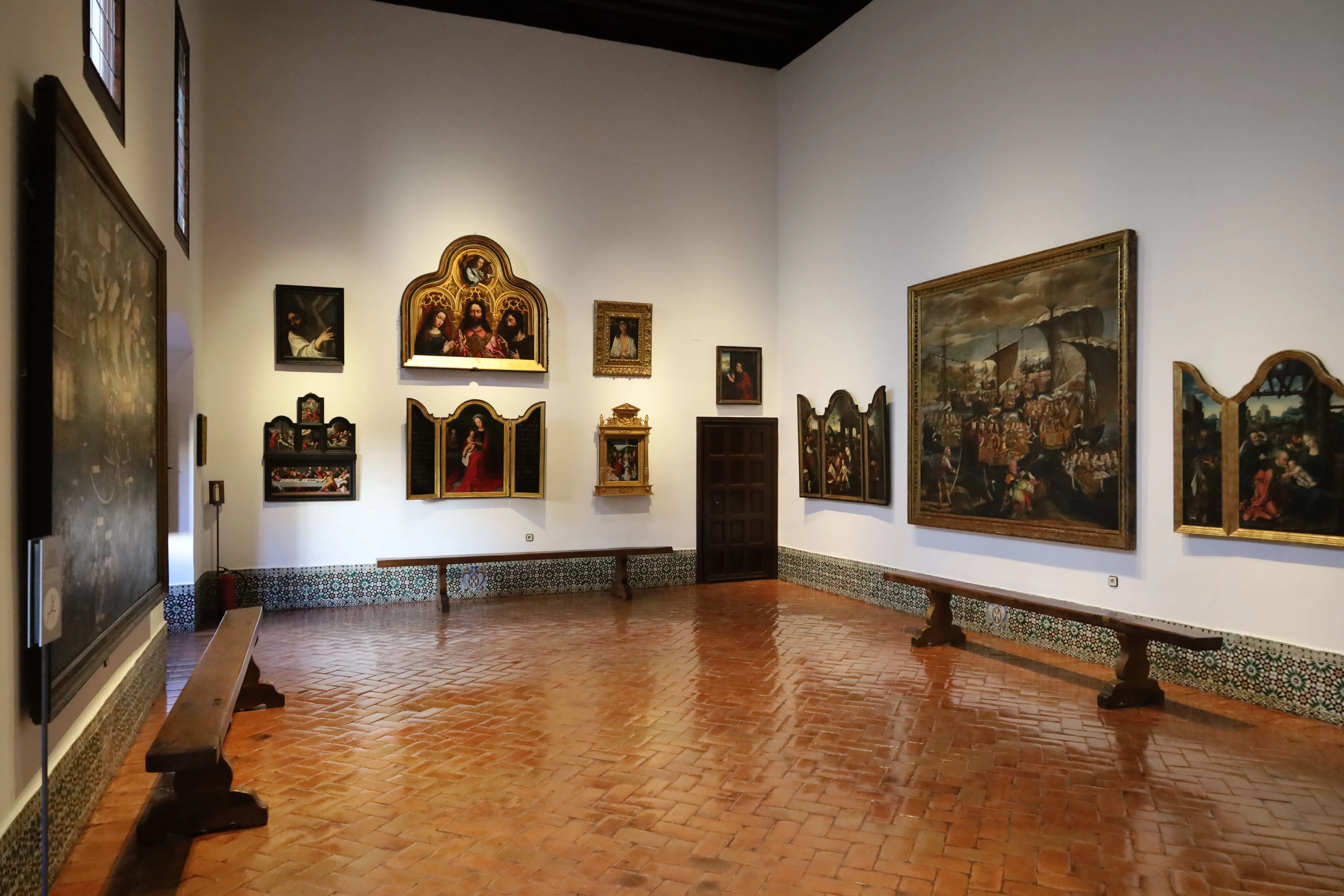Interior gallery with religious paintings and tapestries on white walls, wooden benches, and terracotta floor
