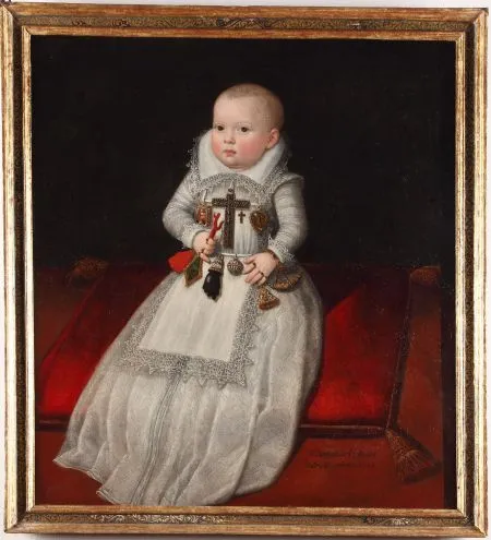 A framed portrait of a young child in a white lace-trimmed dress, holding a cross and other objects, seated on a red cushion against a dark background