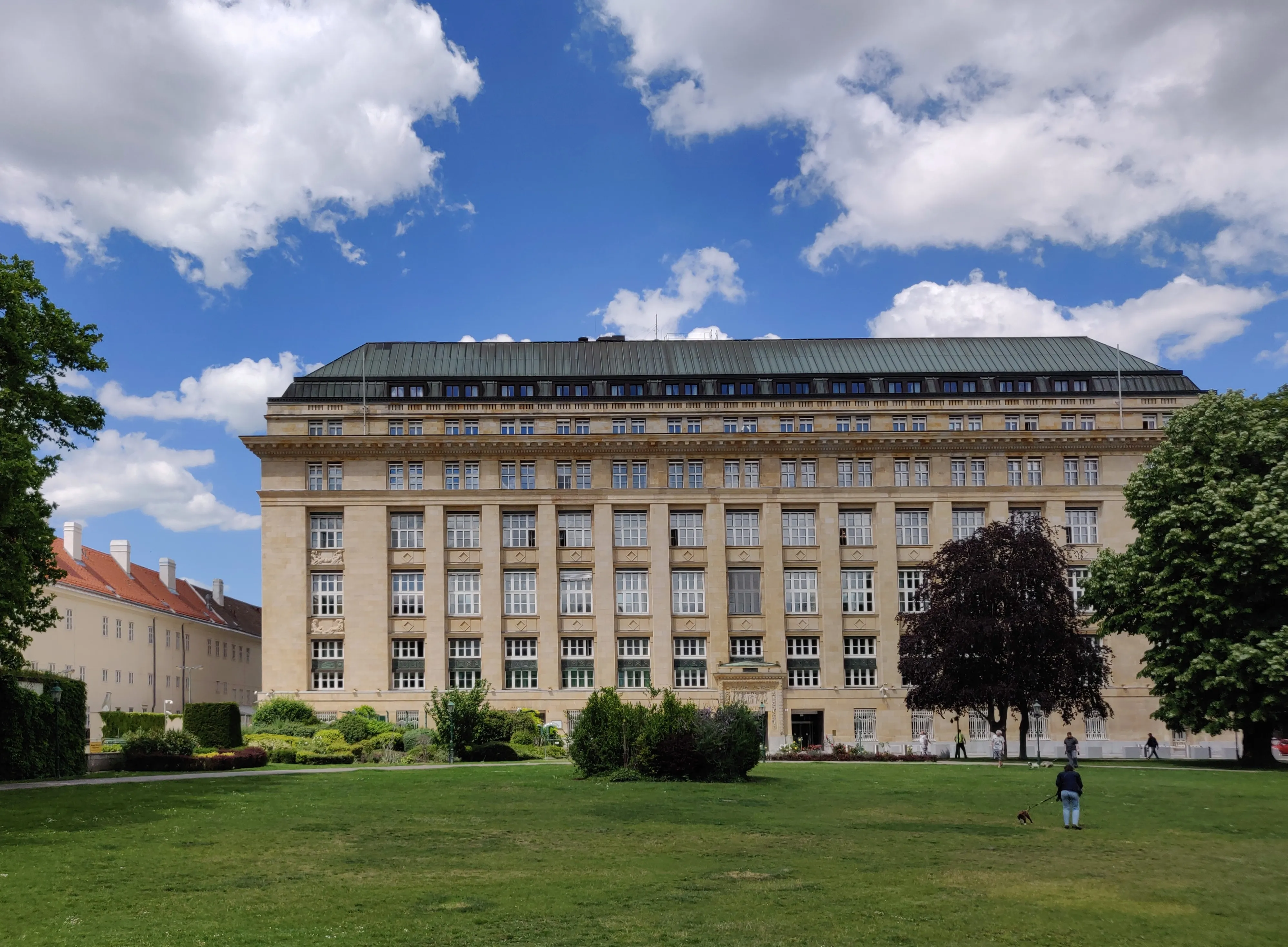 Duży budynek neoklasycystyczny z symetryczną fasadą, licznymi oknami i zielonym dachem, otoczony trawą i drzewami pod błękitnym niebem z białymi chmurami
