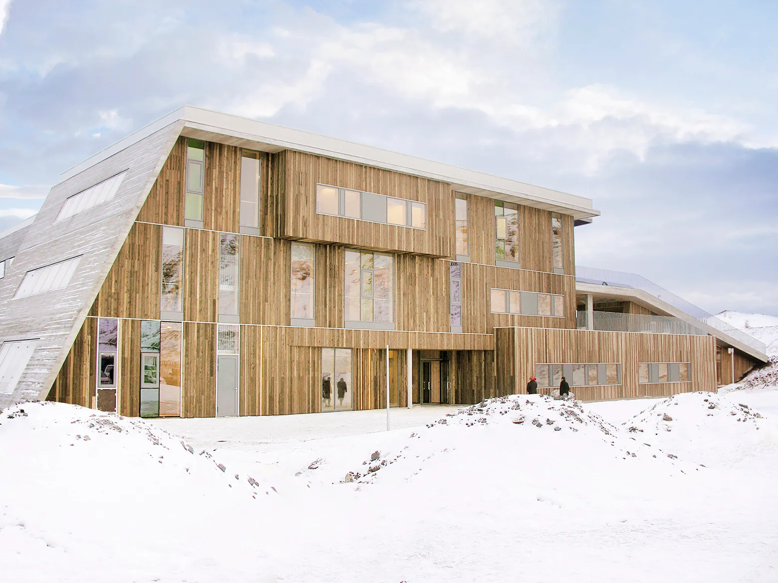 Un edificio moderno revestido de madera con líneas de techo angulares y grandes ventanales rectangulares situado en un paisaje nevado bajo un cielo nublado.