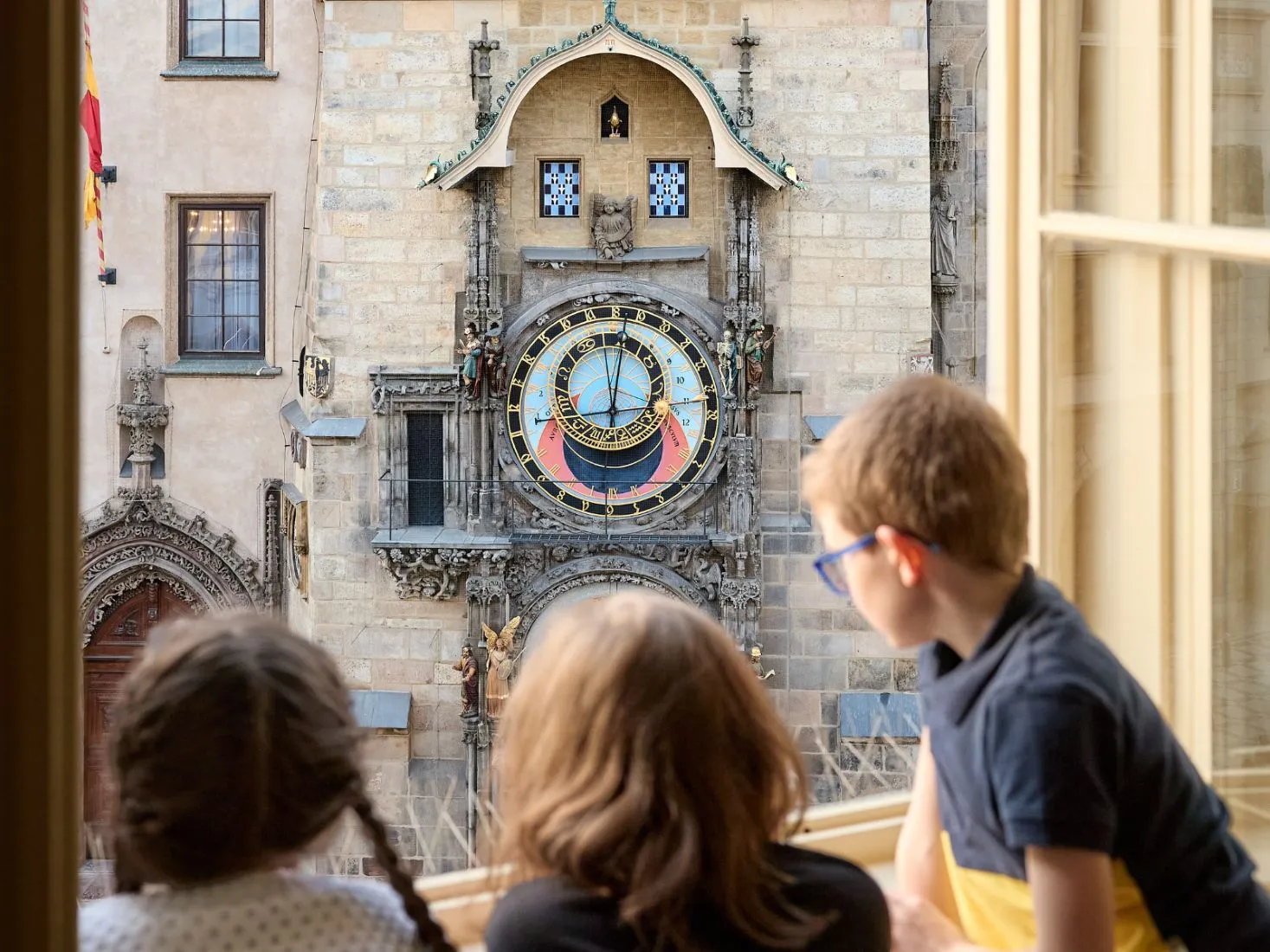 Drie kinderen kijken uit een raam naar de Praagse astronomische klok aan de gevel van een stenen gebouw