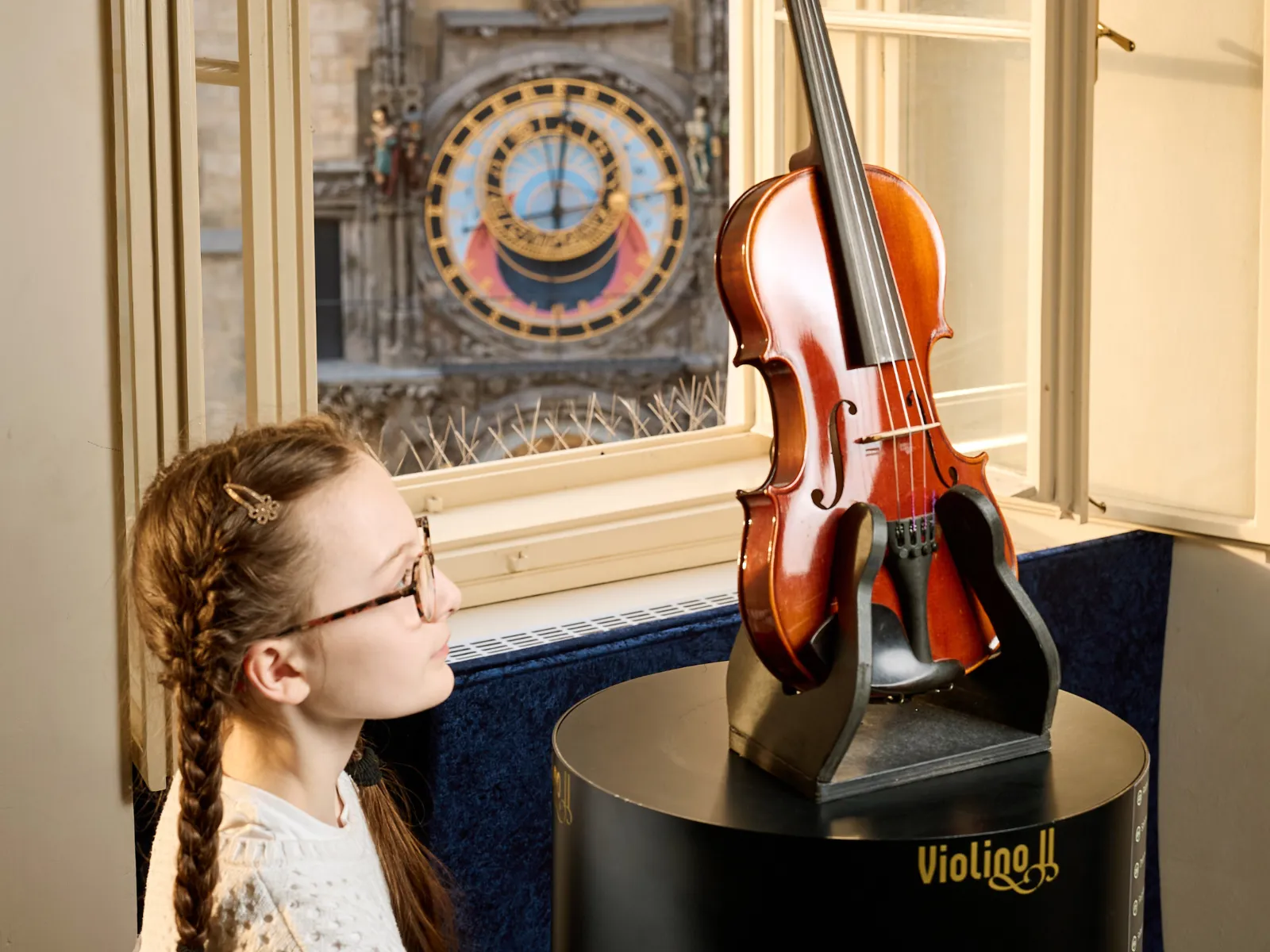 Una niña con gafas y el pelo trenzado mira un violín en exhibición en una muestra de museo con el Reloj Astronómico de Praga visible a través de una ventana.