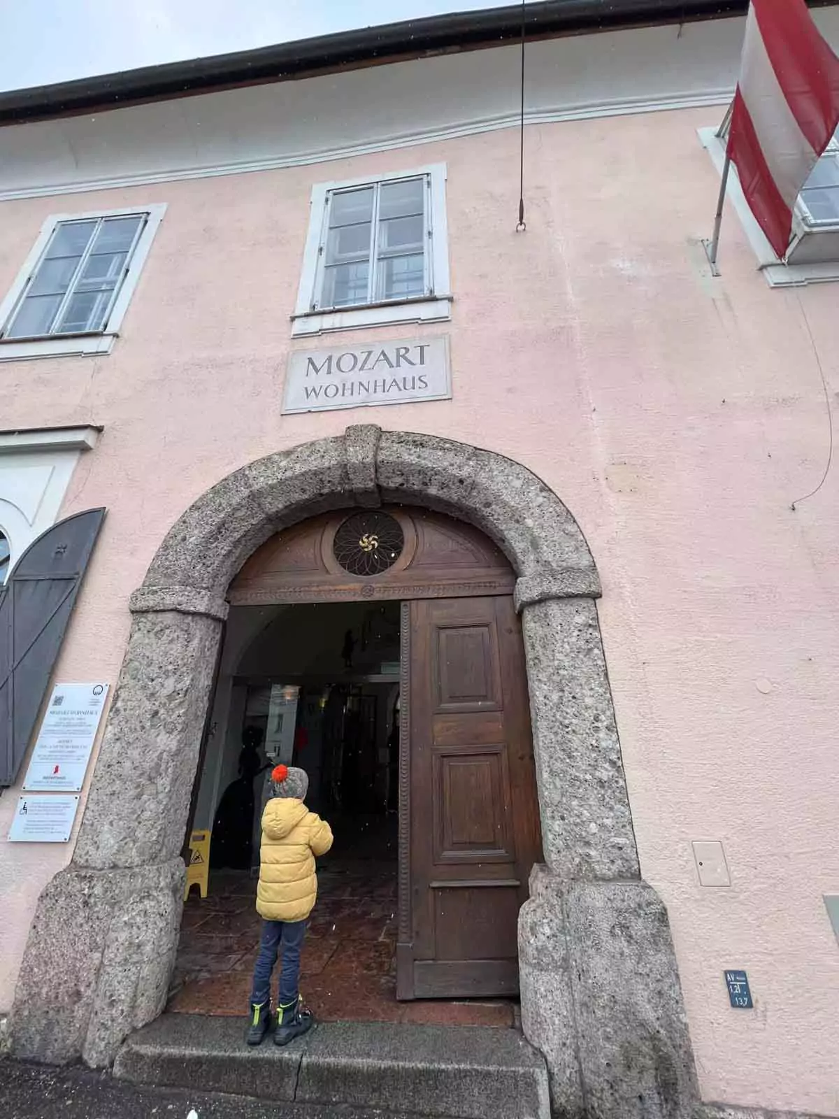 Różowa fasada budynku z kamiennym łukowym wejściem i szyldem z napisem 'Mozart Wohnhaus', z dzieckiem w żółtej kurtce stojącym w drzwiach