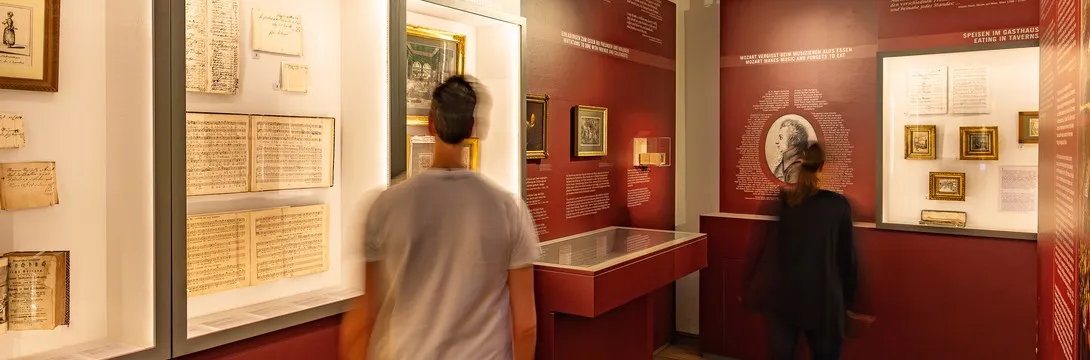 Dos visitantes observando documentos enmarcados y artefactos en vitrinas dentro de la Mozarthaus Vienna.