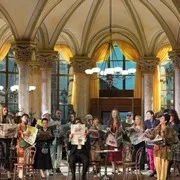 Músicos con vestimenta histórica actuando en un gran salón con ventanas arqueadas y candelabros