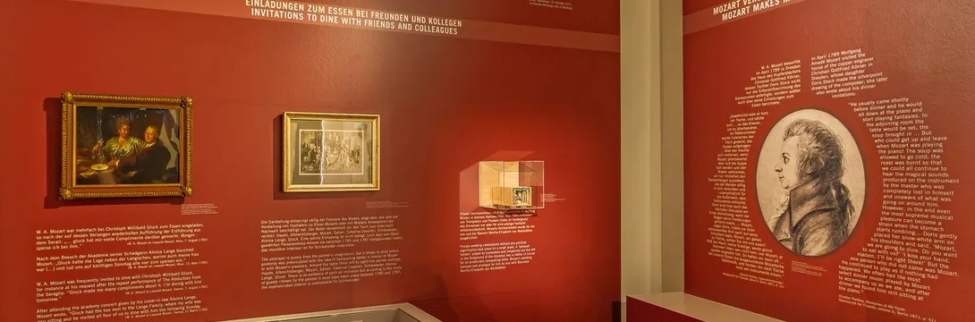 Pared roja de exposición con obras de arte enmarcadas, un retrato circular de Mozart y paneles de texto informativos.