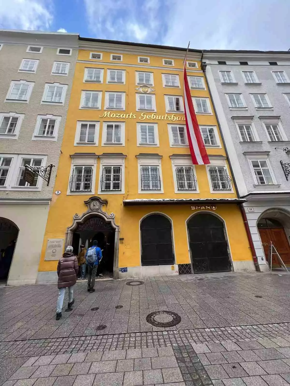 Fachada de edificio amarillo con 'Mozarts Geburtshaus' escrito en letras doradas, una bandera austriaca colgando y gente caminando por la calle adoquinada de abajo.