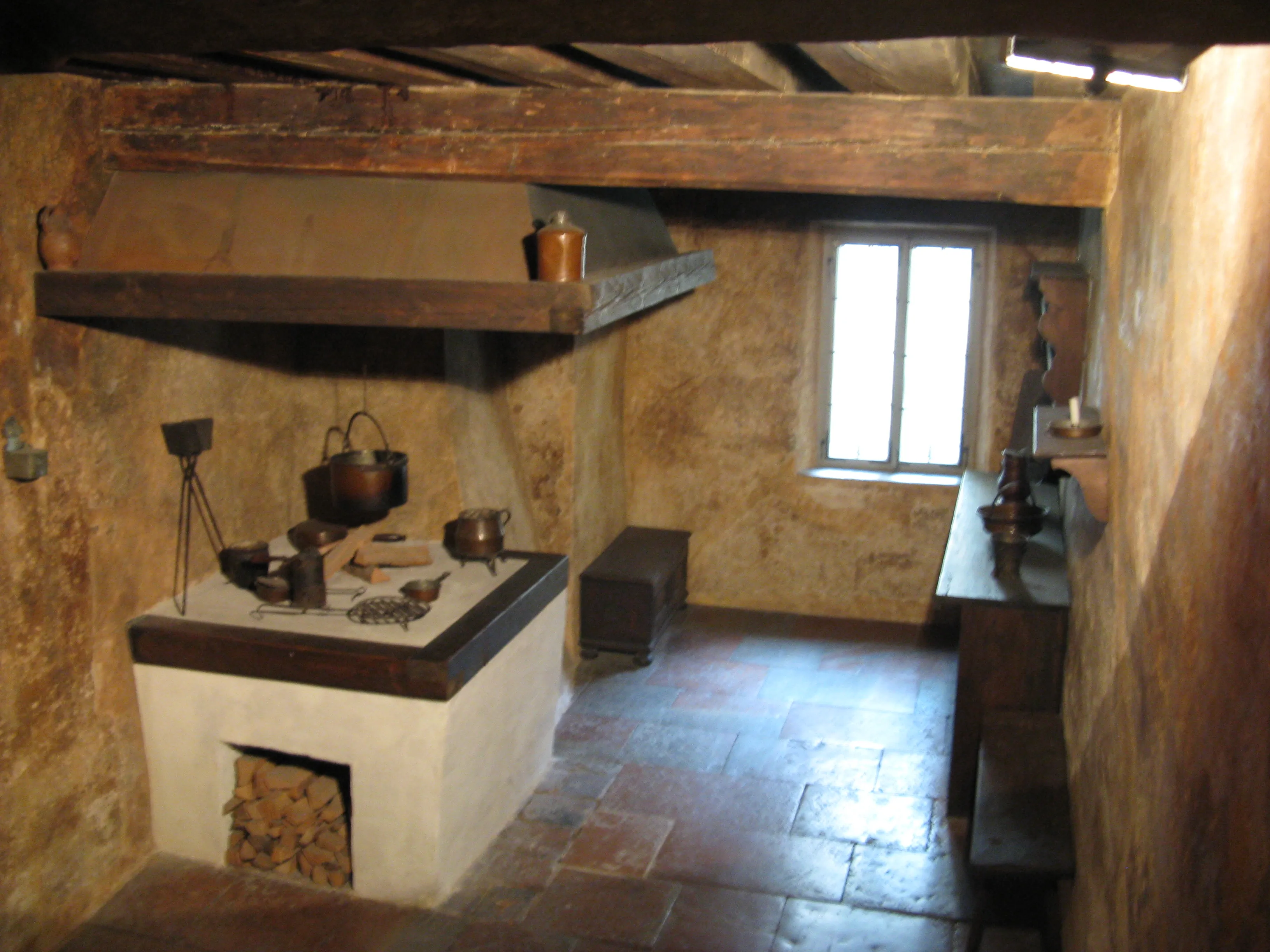 Interior de una cocina histórica que cuenta con una gran estufa de azulejos blancos con leña, vigas de madera en el techo y utensilios de cocina de cobre en exhibición.