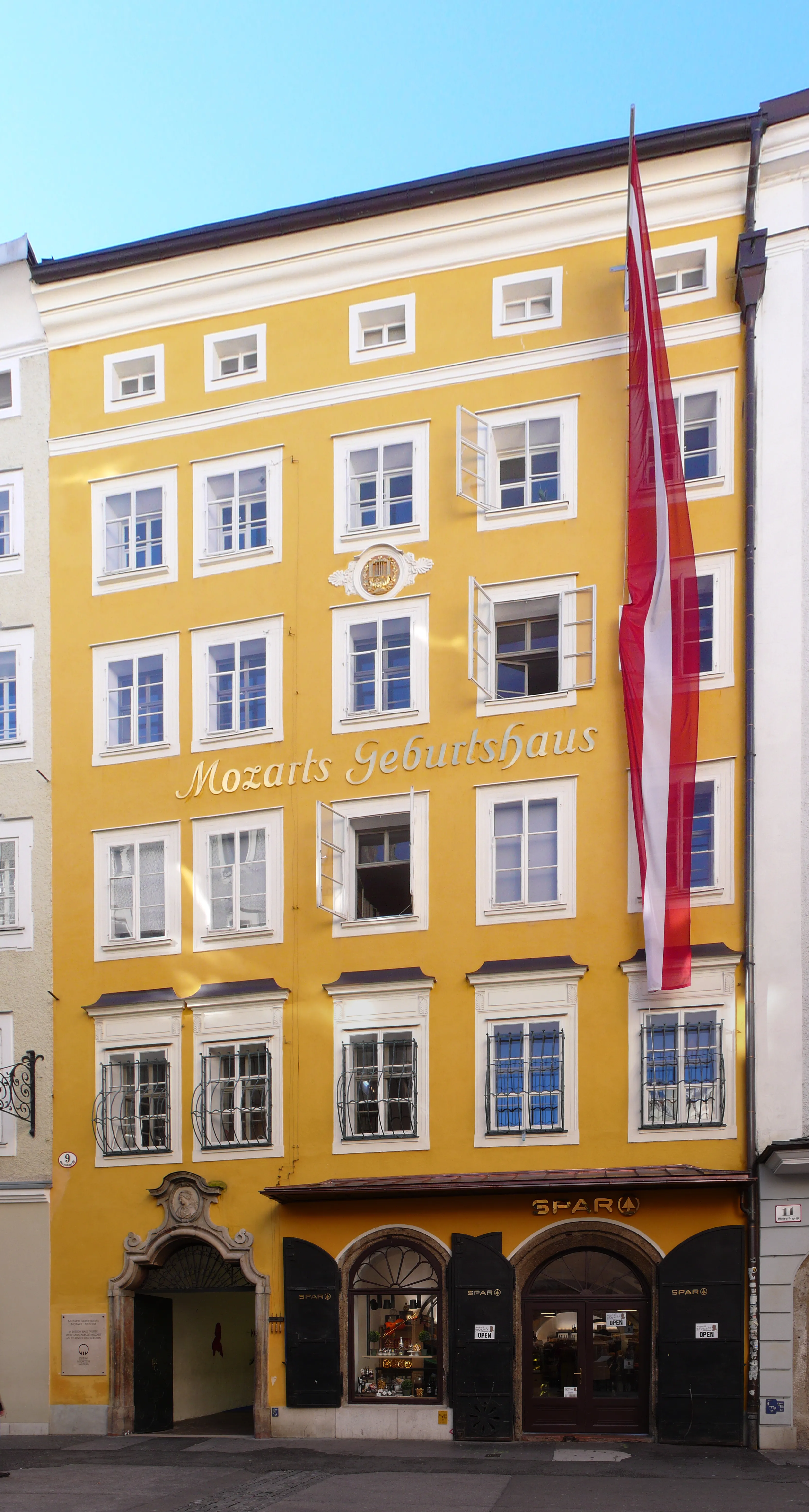Fachada amarilla de un edificio de varias plantas con marcos de ventana blancos, donde se lee 'Mozarts Geburtshaus' en letras doradas, con una gran bandera austriaca colgada verticalmente en el lado derecho.