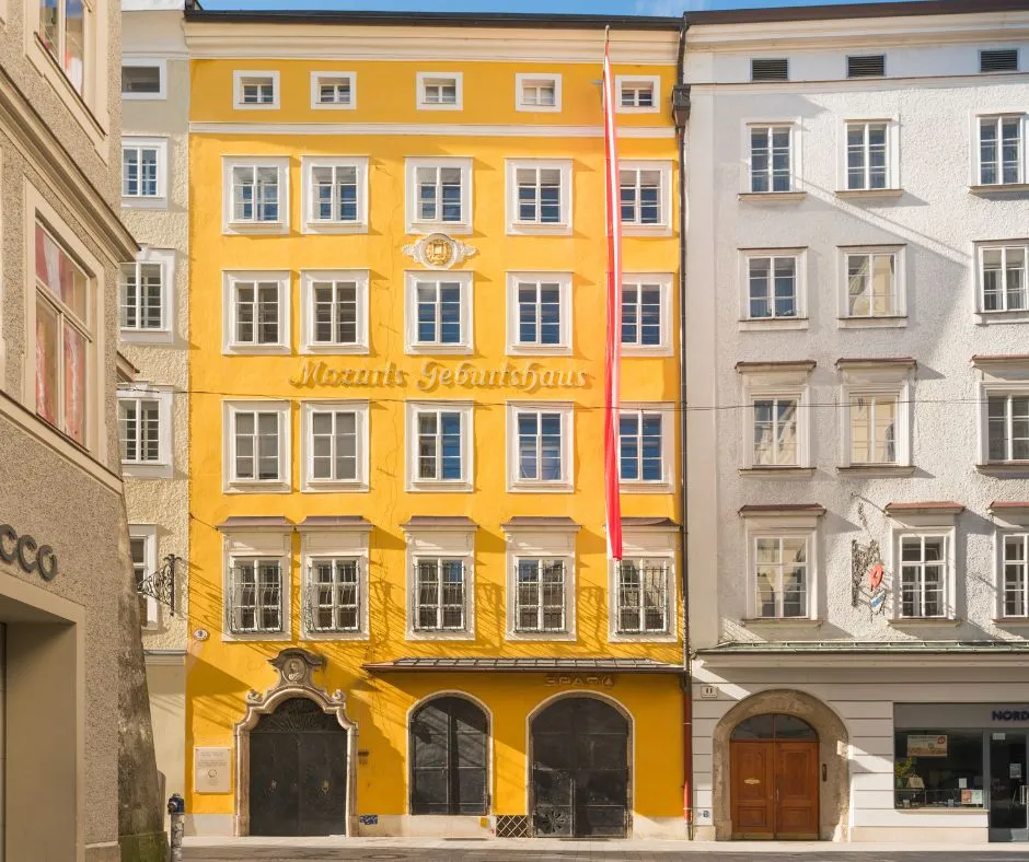 Fachada amarilla de Mozarts Geburtshaus en Getreidegasse con ventanas de marcos blancos, entradas arqueadas en la planta baja y el nombre del edificio inscrito en la pared