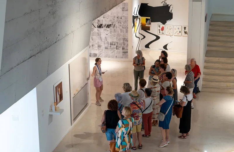 Un grupo de visitantes escucha a un guía en una luminosa galería de arte moderno con grandes obras abstractas en paredes blancas y una escalera a la derecha.