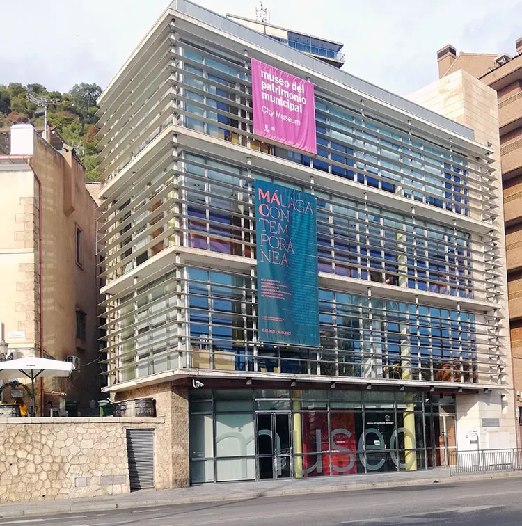 Edificio moderno de fachada de cristal con lamas horizontales, adornado con estandartes rosas y verde azulado que dicen "museo del patrimonio municipal" y "MÁLAGA CONTEMPORÁNEA", con grandes letras de "museo" en los ventanales de la planta baja.