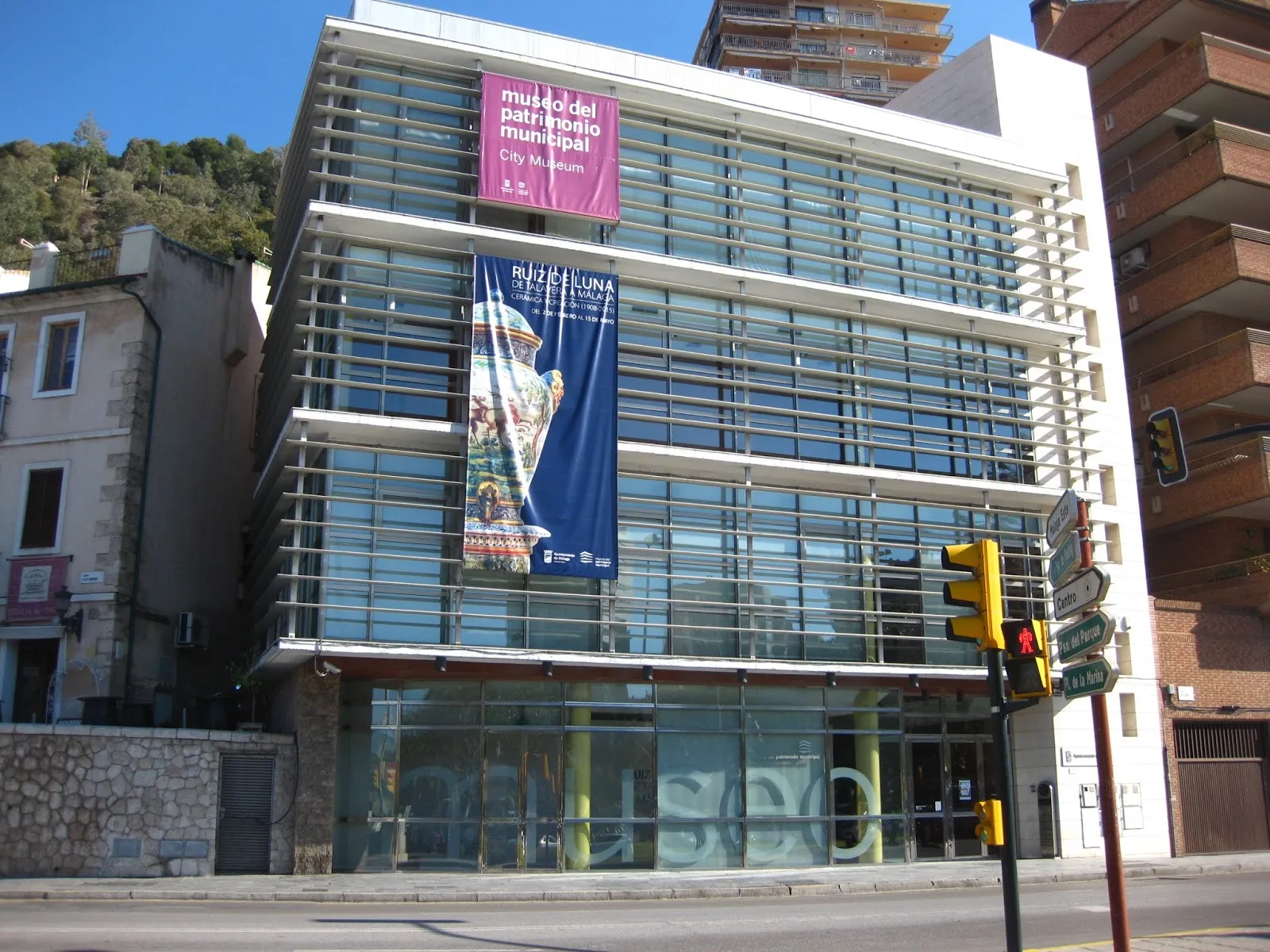 Edificio moderno de varias plantas con fachada de muro cortina de cristal, que luce una pancarta rosa con el texto 'museo del arte moderno y contemporaneo' y una pancarta azul con 'MÁLAGA', visto desde la calle con semáforos en primer plano.