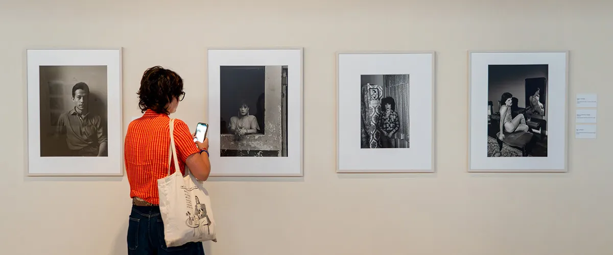 Un visitante con una camisa de rayas naranjas se encuentra frente a cuatro retratos fotográficos en blanco y negro enmarcados y expuestos en una pared blanca de la galería.
