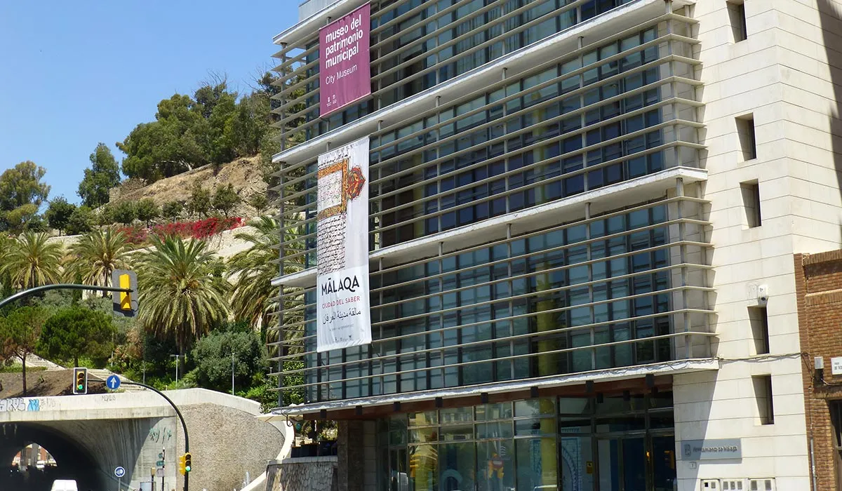 Moderno edificio de museo con fachada de cristal y carteles de exposición verticales, con una colina con palmeras de fondo y una entrada de túnel visible a nivel de calle