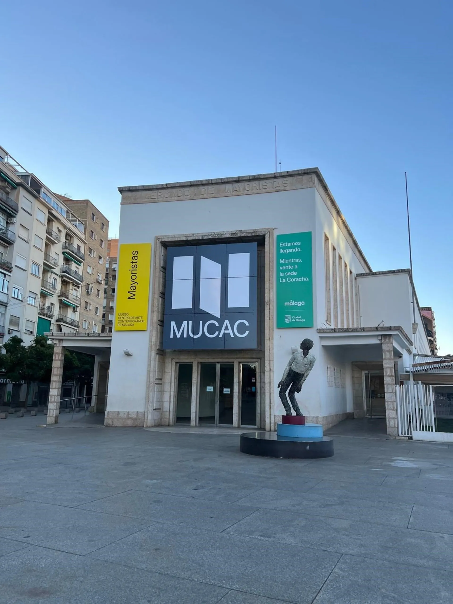 Vista exterior del edificio del MUCAC con un cartel grande con el nombre del museo, pancartas amarillas y verdes, y una escultura de una figura sobre un pedestal en la plaza.