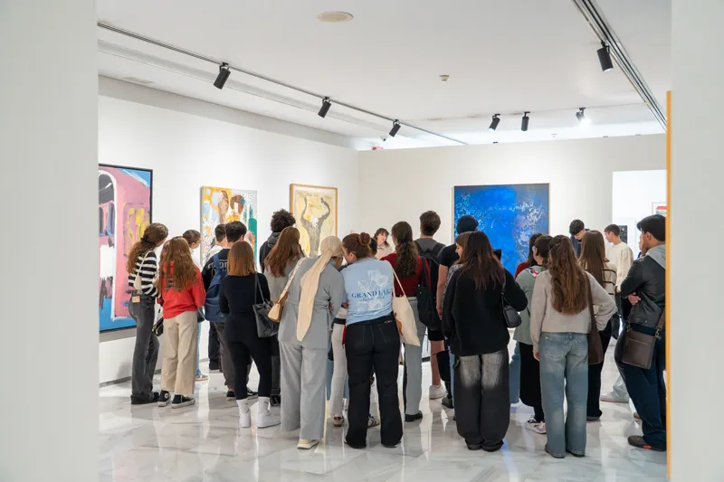Un grupo de visitantes se reunió en una galería de paredes blancas y luminosas para contemplar cuadros enmarcados bajo la iluminación de focos de techo.