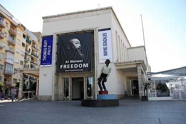 Vista exterior del edificio MUCAC La Coracha con una gran pancarta de Ai Weiwei Freedom en la fachada y una escultura frente a la entrada