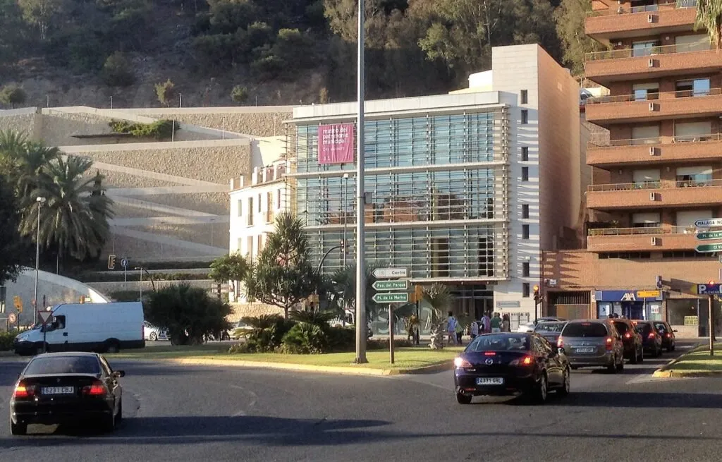 Edificio moderno de fachada de cristal con un letrero rosa de MUCAC, visto desde una calle con coches y una ladera al fondo