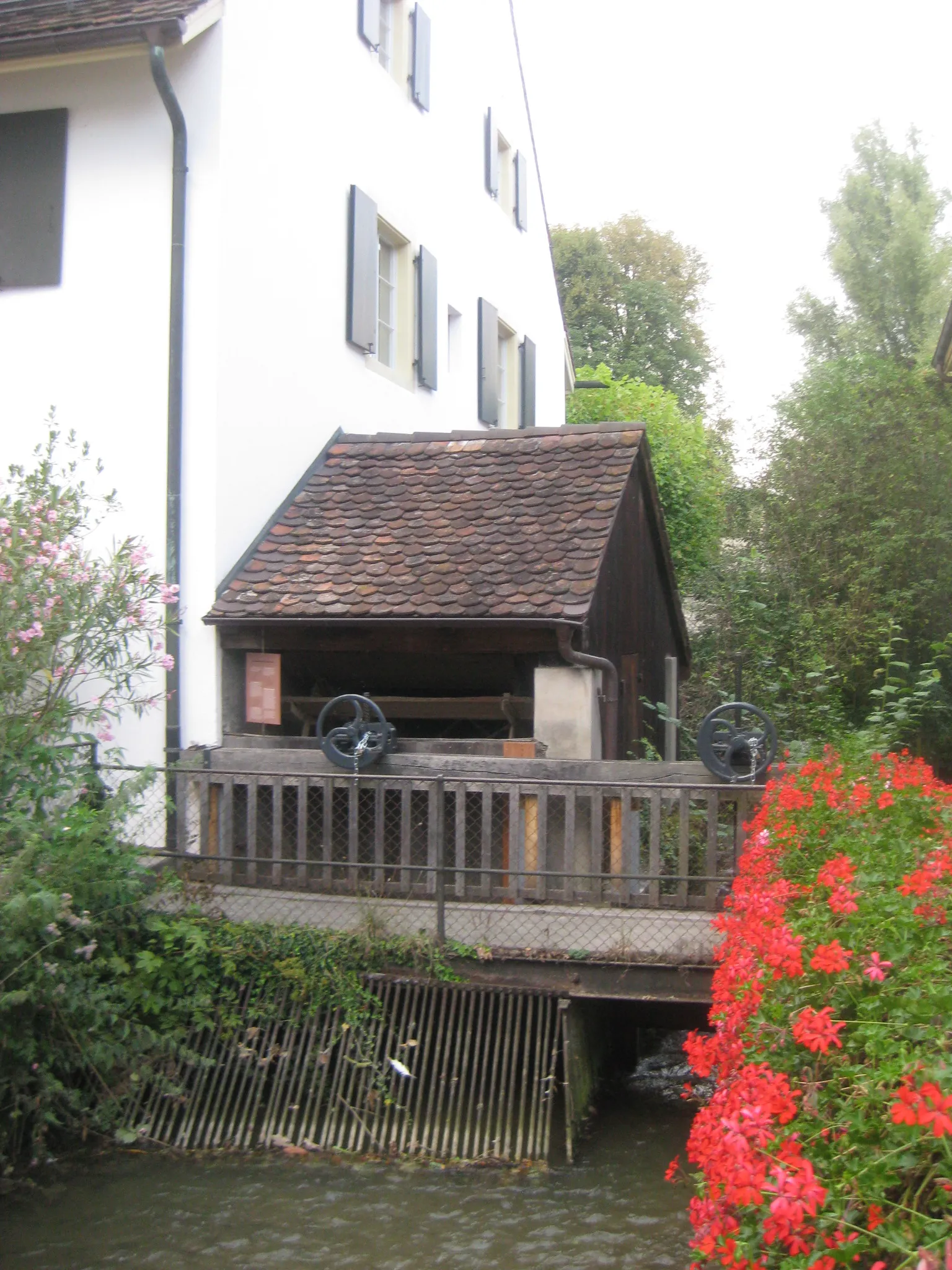 Widok z zewnątrz na młyn wodny Mühlemuseum Brüglingen, przedstawiający biały budynek z ciemnymi okiennicami, drewnianą obudowę koła wodnego z dachem krytym dachówką, kratę wlotową wody oraz czerwone kwiaty wzdłuż strumienia.