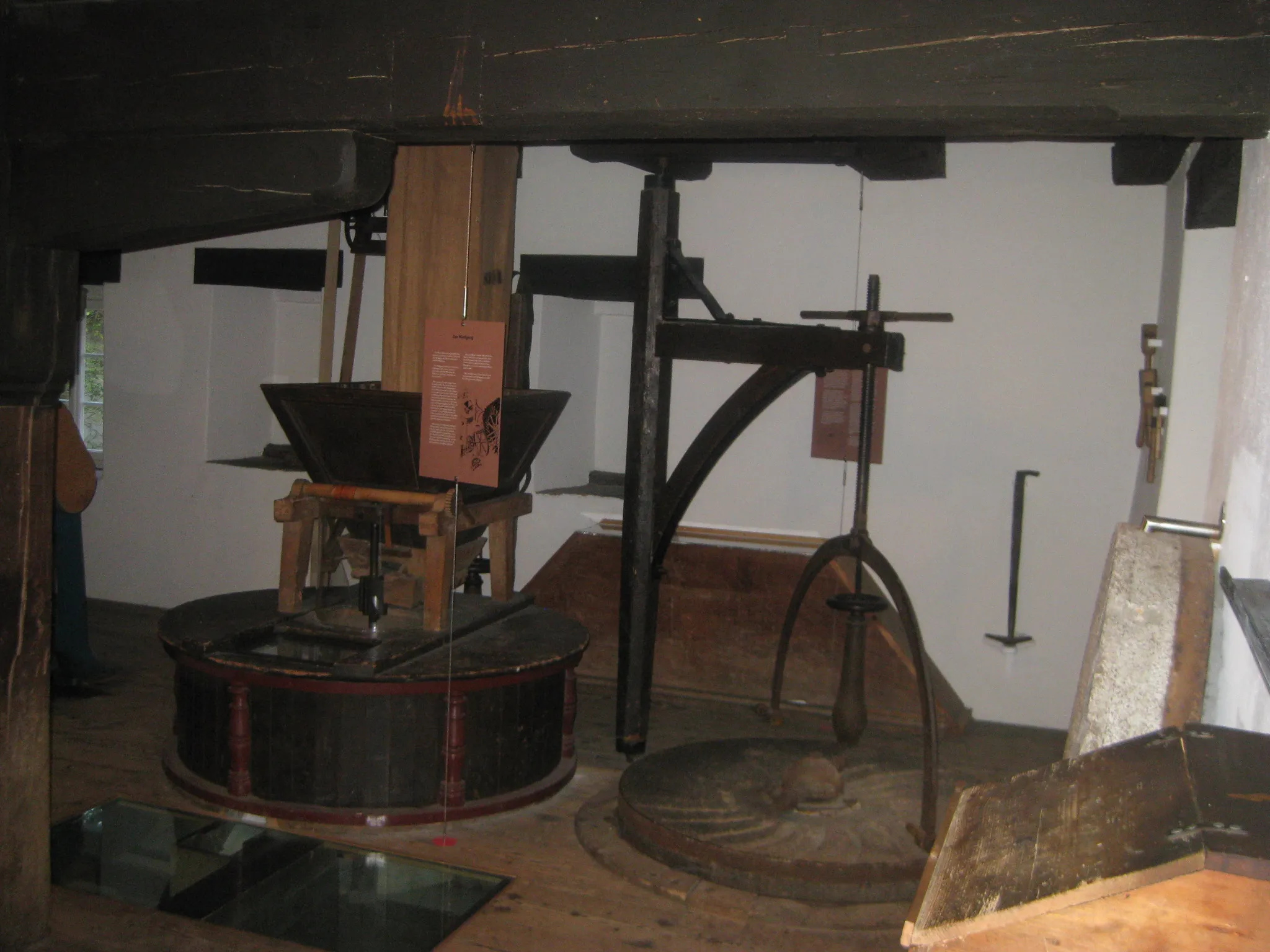 Wnętrze Mühlemuseum Brüglingen prezentujące zabytkowe maszyny młyńskie, w tym duży mechanizm koła drewnianego, metalowy zasobnik oraz kamienie młyńskie pod ciemnymi drewnianymi belkami stropowymi z przymocowanymi tablicami informacyjnymi.