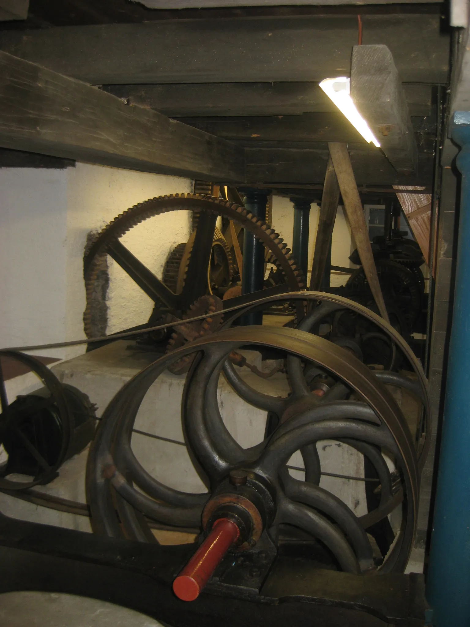 Duże drewniane i metalowe koła zębate, szprychowe koła zamachowe oraz skórzane pasy napędowe zabytkowych mechanizmów młyna wodnego wewnątrz muzeum, z korbą z czerwoną rączką na pierwszym planie i drewnianymi belkami stropowymi powyżej.