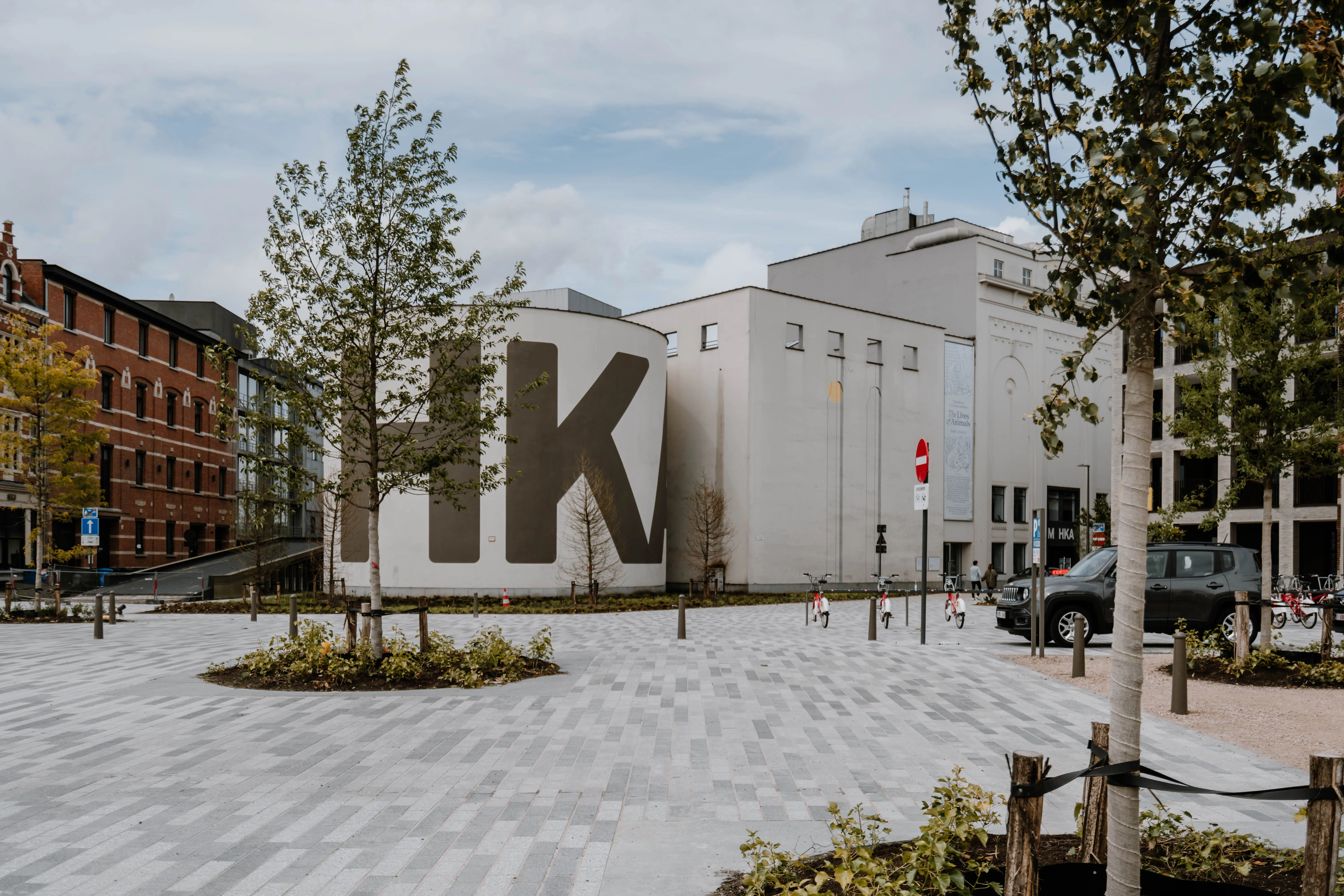 Vista exterior del edificio del museo M HKA con grandes letras HK pintadas en la fachada blanca de la antigua nave de grano, rodeado por una plaza pavimentada con árboles.