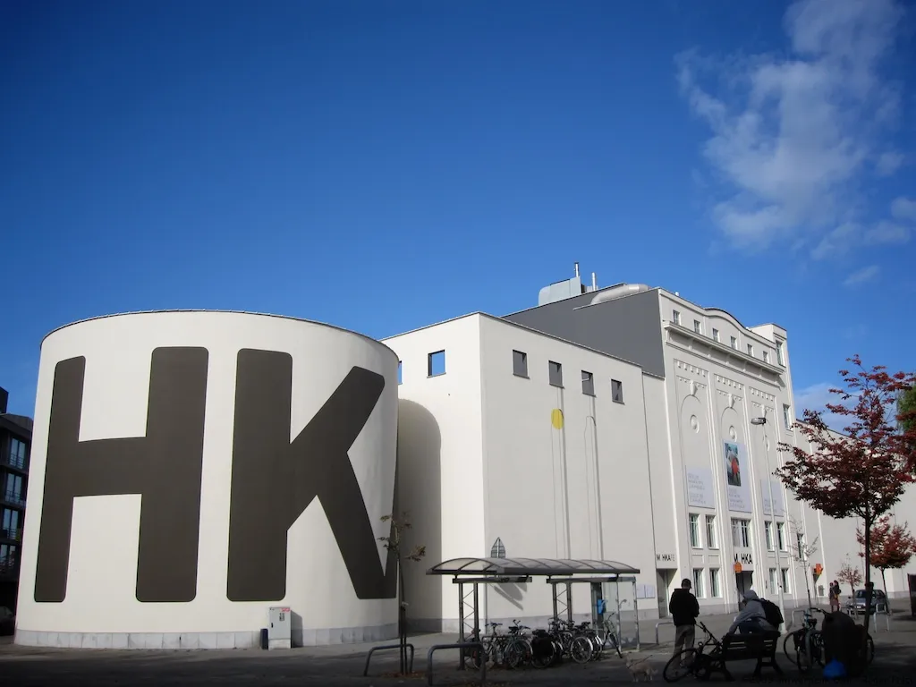 Biała cylindryczna konstrukcja silosu na zboże z dużymi czarnymi literami HK obok budynku muzeum M HKA pod błękitnym niebem