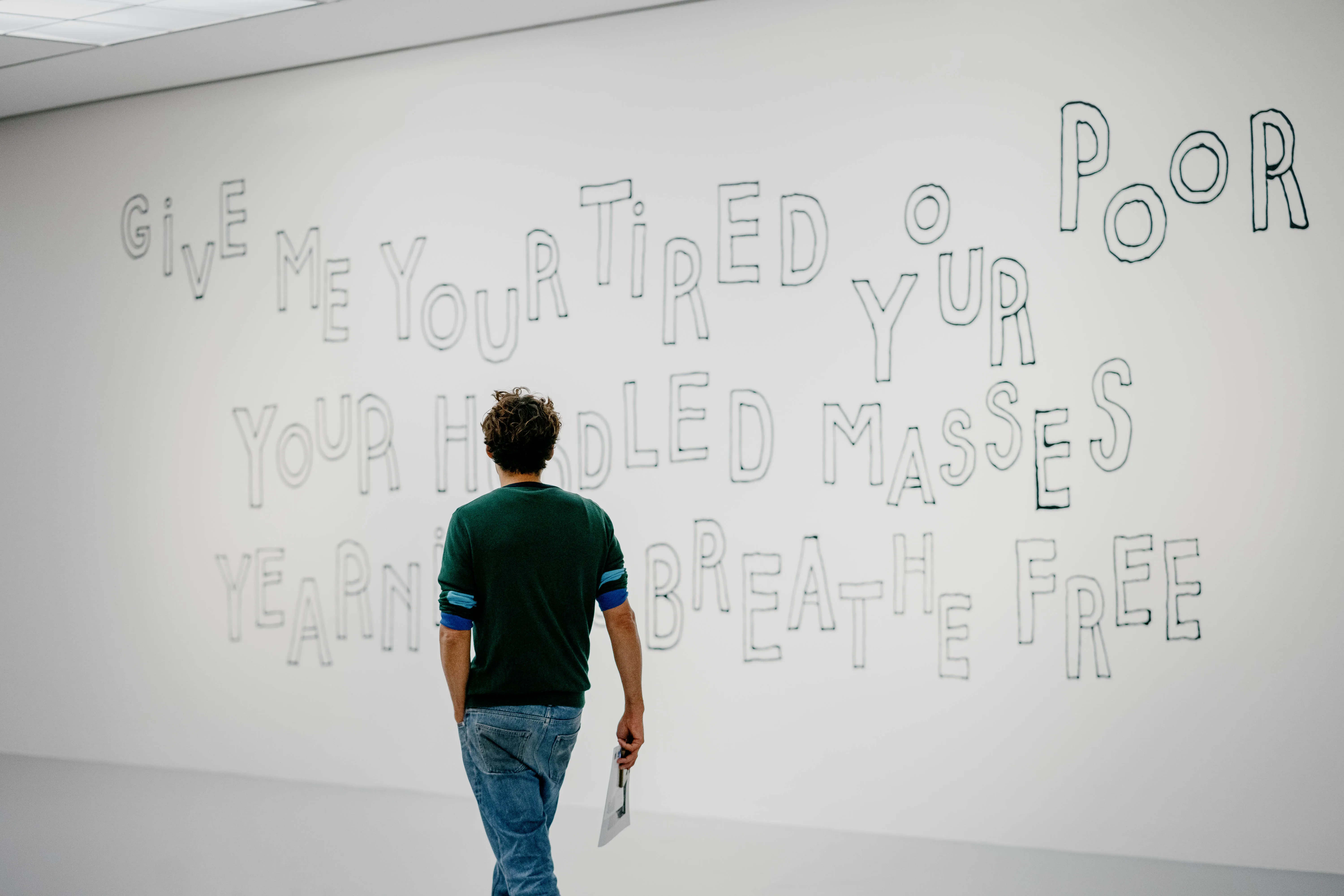 Un visitante se encuentra ante una gran pared blanca de una galería que muestra un texto perfilado a mano que reza: 'Give me your tired your poor your huddled masses yearning to breathe free'