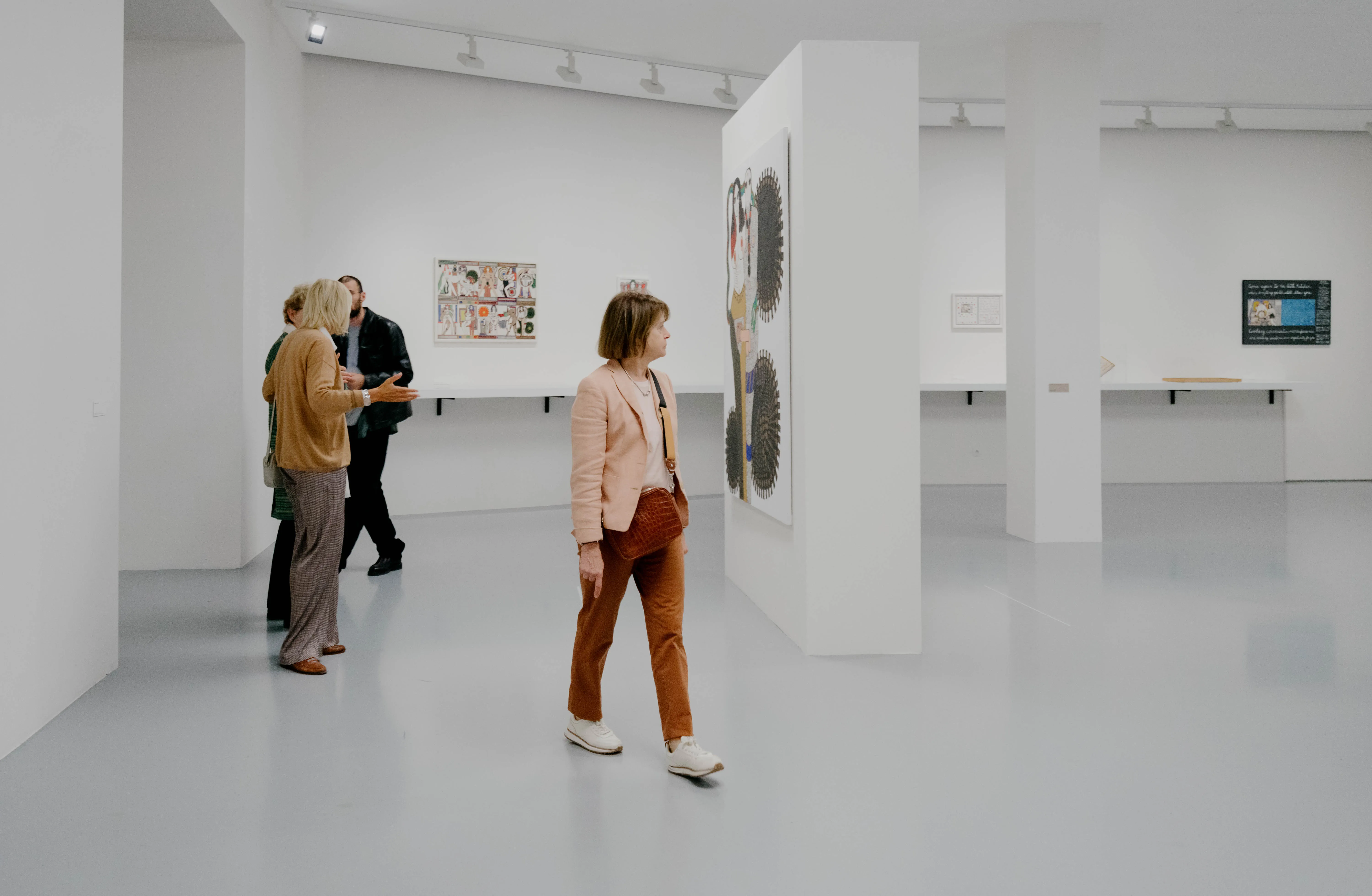 Visitantes caminando a través de una galería de museo blanca y luminosa, con obras de arte expuestas en las paredes y un tabique central.