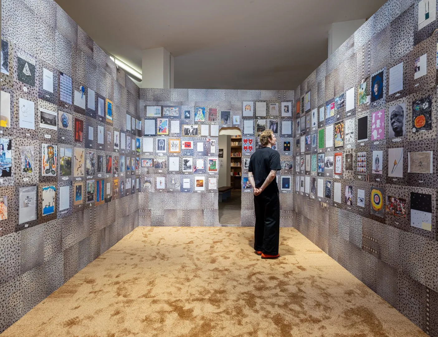 Sala de museo con las paredes cubiertas por una densa cuadrícula de pequeñas obras de arte y grabados enmarcados, con una persona vestida de oscuro de pie en el centro observando la exposición.