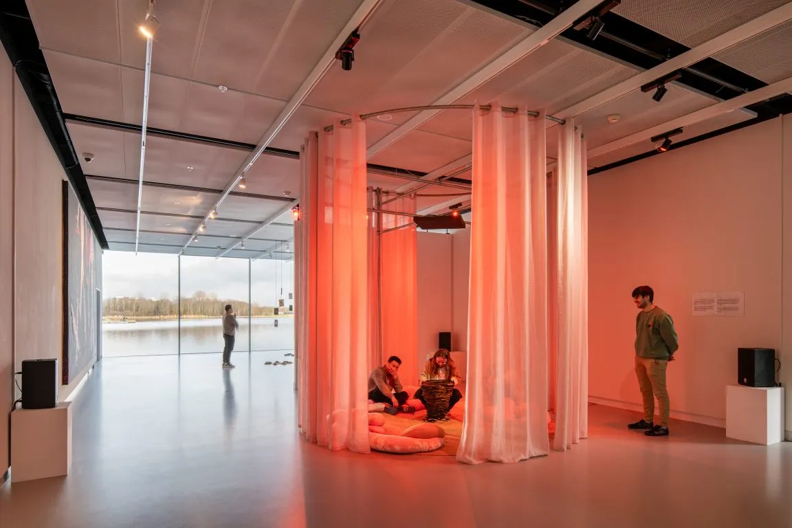 Interior de una galería de museo que presenta una gran instalación circular de tela rosa con personas sentadas en su interior y de pie cerca, con grandes ventanales a lo largo de la pared izquierda que muestran una vista al exterior.