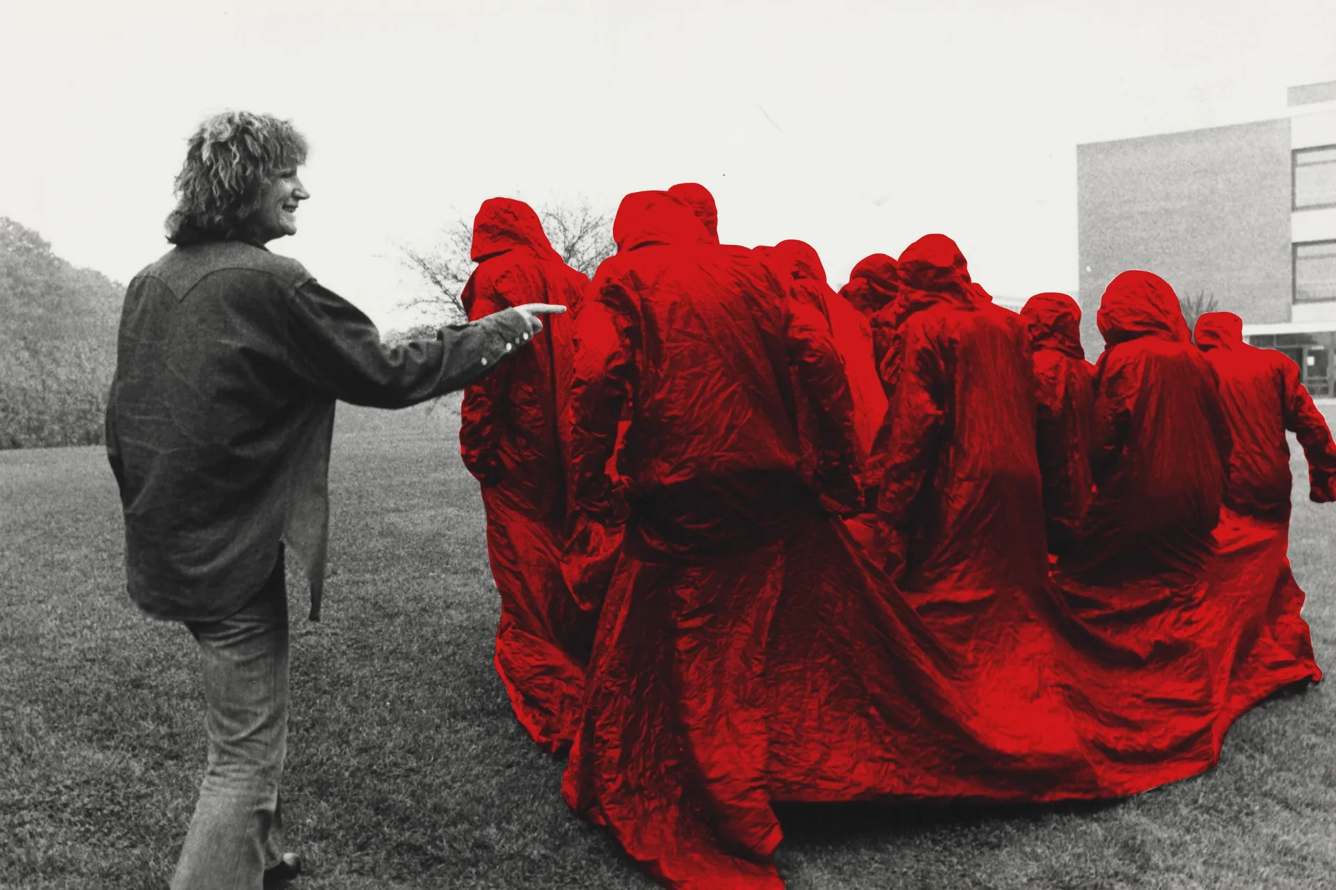 Una persona con chaqueta oscura señala a un grupo de figuras con túnicas encapuchadas de color rojo brillante reunidas al aire libre sobre el césped, con un enfoque de color selectivo que resalta el rojo sobre un fondo en blanco y negro.