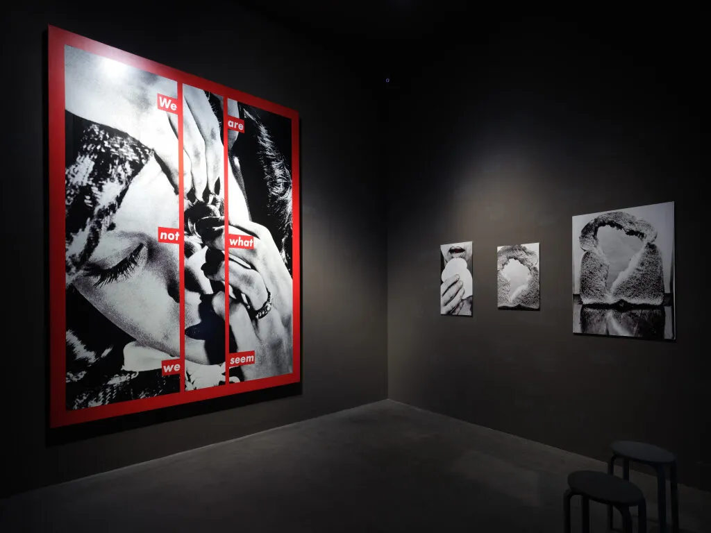 Sala de galería oscura que presenta grandes fotografías en blanco y negro, incluyendo una obra destacada con marco rojo que muestra un rostro y texto, además de impresiones más pequeñas enmarcadas en la pared contigua.
