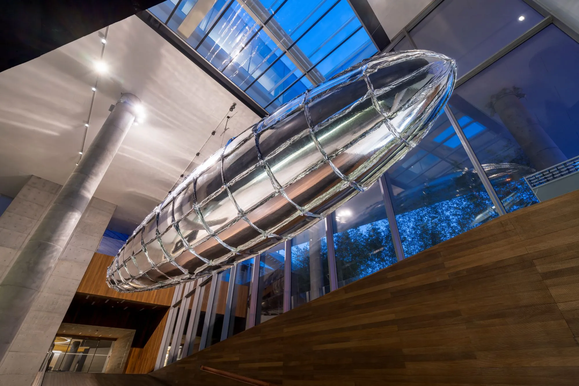 Gran escultura plateada reflectante con forma de zeppelín suspendida del techo dentro de un edificio moderno con paredes de cristal y paneles de madera.
