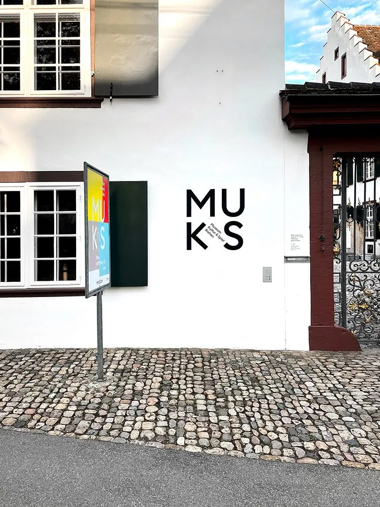Fachada blanca con el logotipo de MUKS en la pared, patio empedrado y puerta de entrada con detalles de hierro forjado