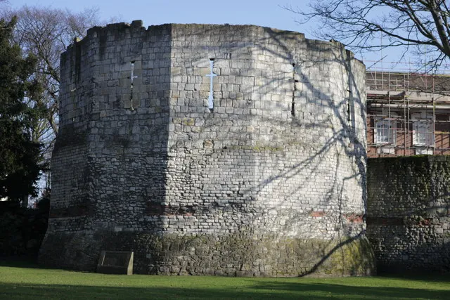 Rounded stone tower with narrow slit windows standing on a grass lawn, tree shadows cast across the weathered masonry, with scaffolding visible on the right side
