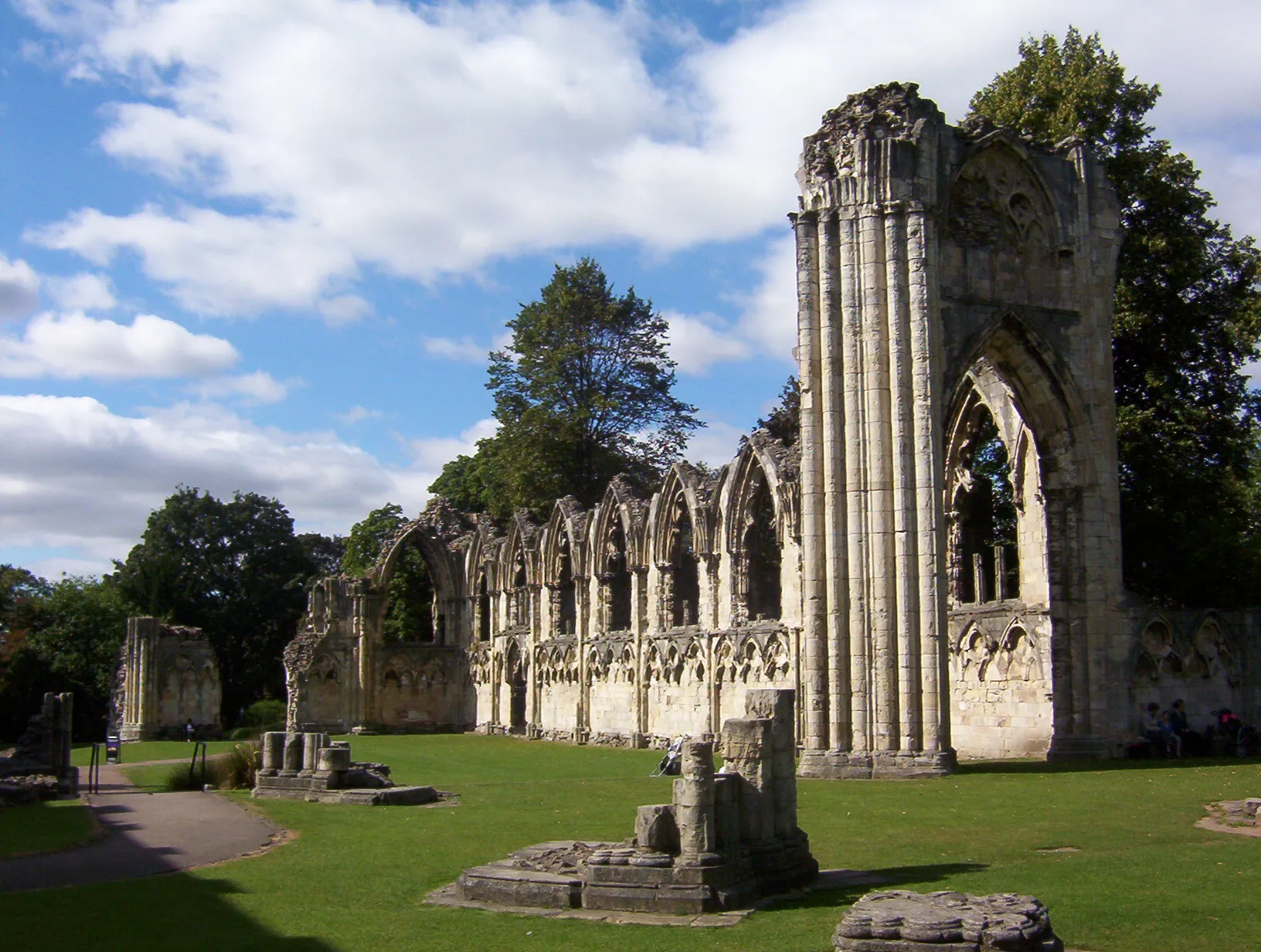 Ruins of St Mary's Abbey with tall Gothic stone arches and columns standing on green lawns under a blue sky with white clouds