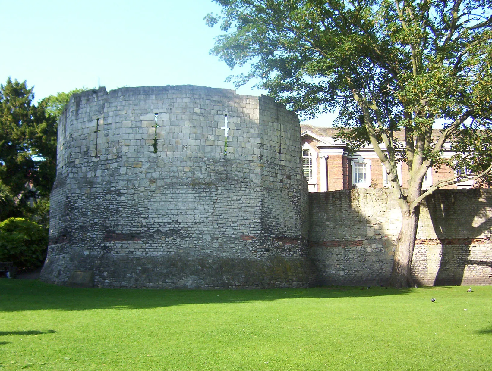 Rounded stone Multangular Tower with narrow slit windows standing on a green lawn with trees and a brick building in the background