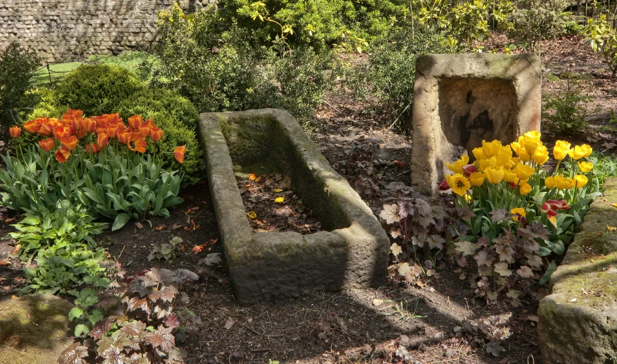 Stone trough and carved niche set in a garden bed surrounded by orange tulips and yellow daffodils.