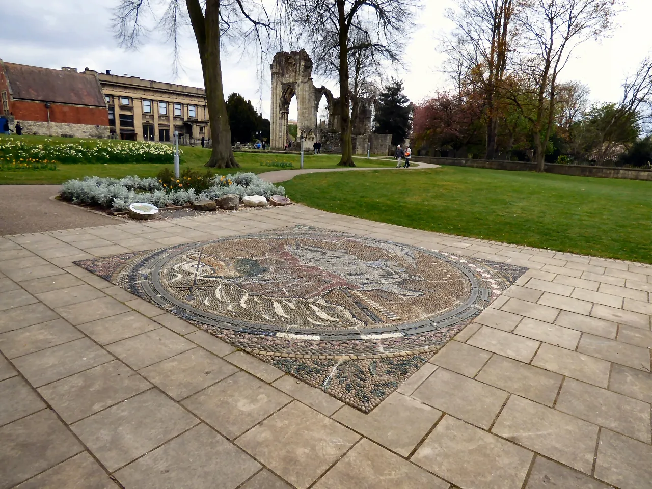 Circular geological map mosaic set into the paved walkway of York Museum Gardens, with St Mary's Abbey ruins and the Yorkshire Museum building in the background